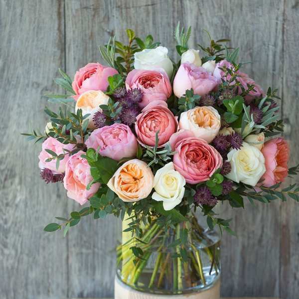 Arrangement of pink, peach, and white roses with purple accents in a clear glass vase