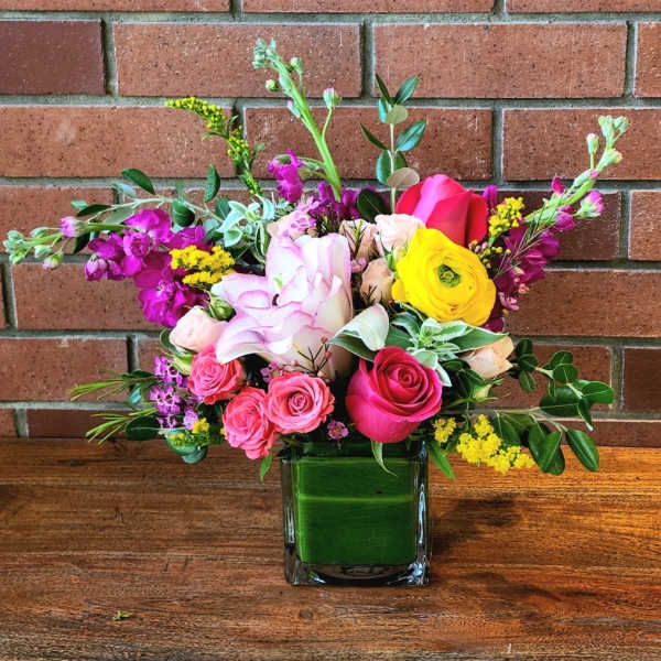 Bright mixed bouquet of pink roses and yellow ranunculus in a square glass vase lined with green leaves