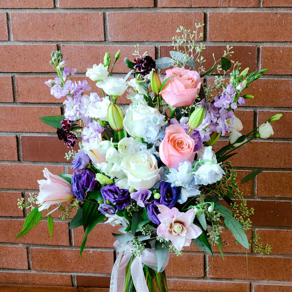 Tall bouquet of pink roses, purple lisianthus, and lilies in a clear glass vase with ribbon