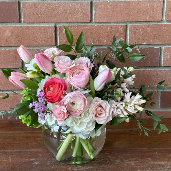 Low glass vase of pink tulips, ranunculus, roses, and white blooms in a rounded bouquet.