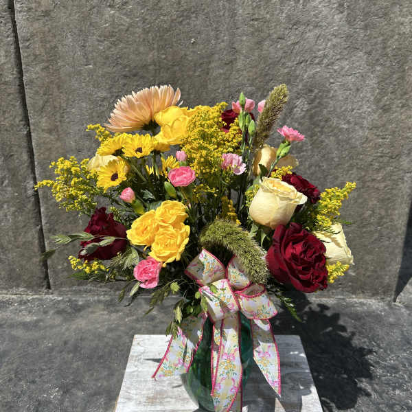 Mixed bouquet of yellow, pink, red, and cream flowers in a glass vase with a ribbon