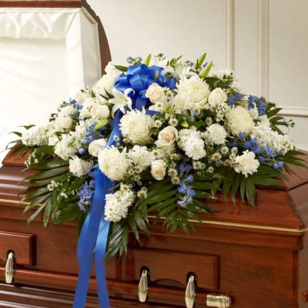 White and blue funeral casket spray with roses, lilies, and mums accented by a blue ribbon on a wood casket