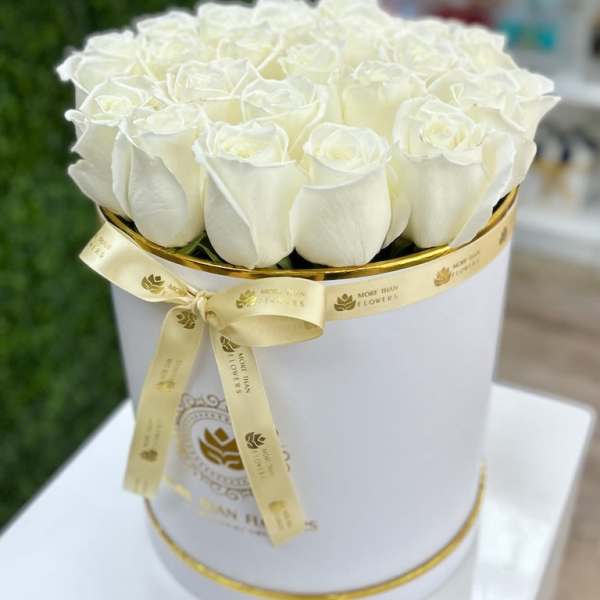 Round white hatbox filled with white roses and wrapped with a gold logo ribbon.
