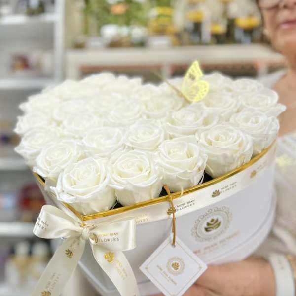 Heart-shaped white box filled with white roses, tied with a white ribbon and tag.