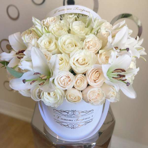 Round hatbox arrangement of cream roses and white lilies in a white container with script text
