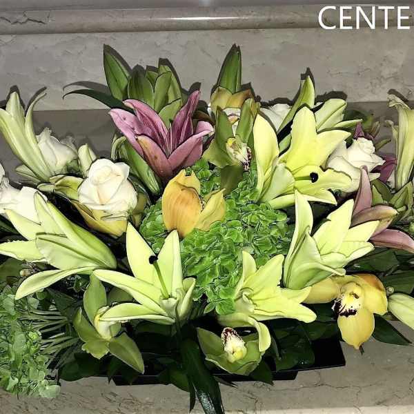 Low centerpiece of white roses, cream lilies, green hydrangea, and orchids in a rectangular black container