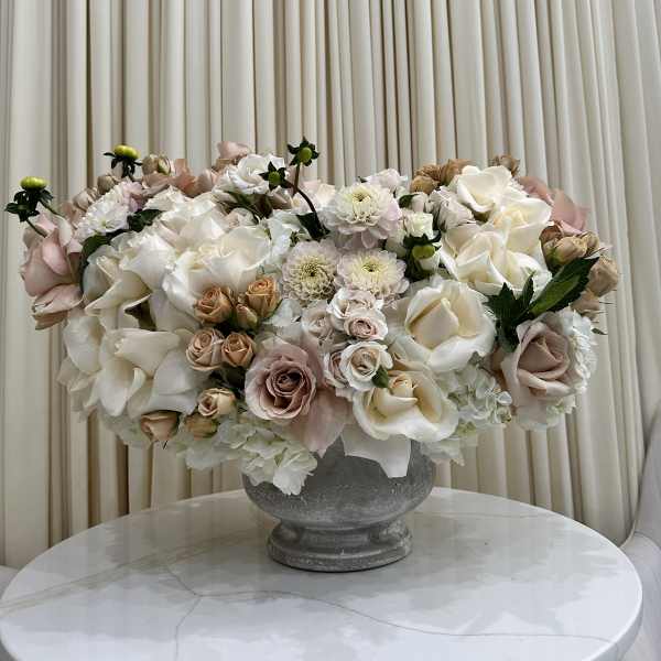 Low centerpiece of ivory and blush roses and white blooms in a stone compote vase on a round table