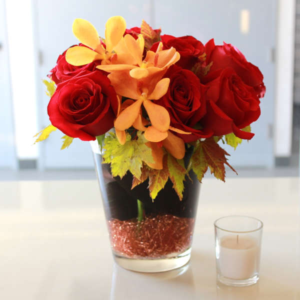 Red roses and orange flowers in a glass vase beside a small candle