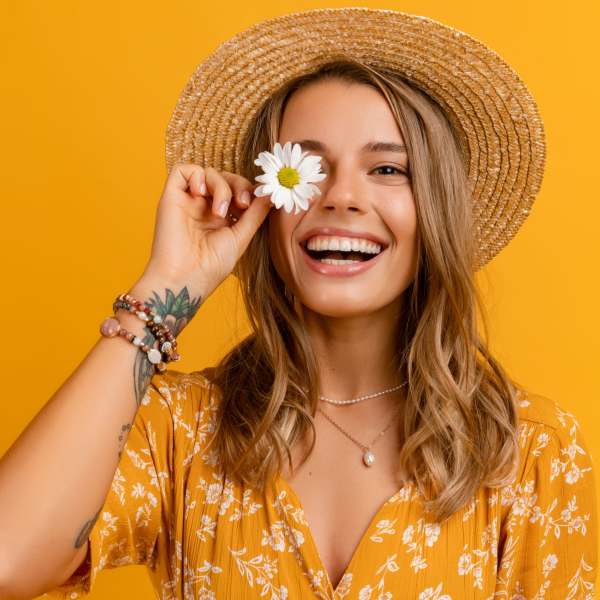 Smiling woman in a straw hat holding a white daisy over one eye