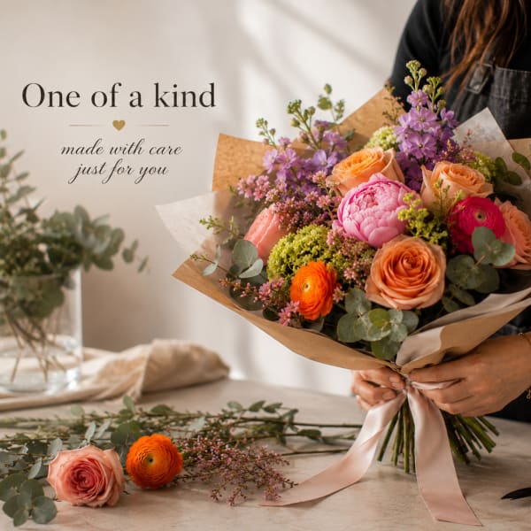 Hand-tied bouquet of peach, pink, and purple flowers wrapped in paper