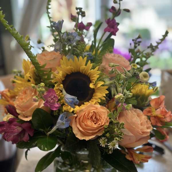 Mixed bouquet with a sunflower, peach roses, and purple blooms in a glass vase