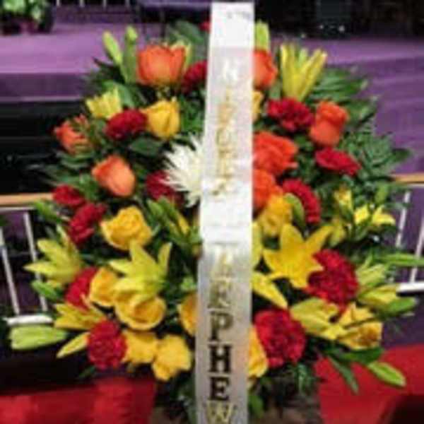Standing funeral spray of red, yellow, and orange flowers with a white ribbon
