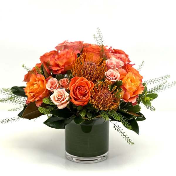 Low orange and peach rose arrangement with pincushion protea in a glass cylinder vase.