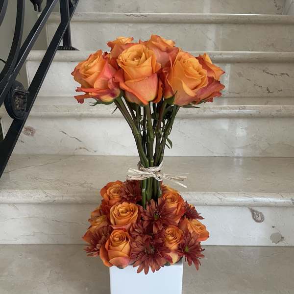 Orange roses and burgundy daisies arranged in a white square vase