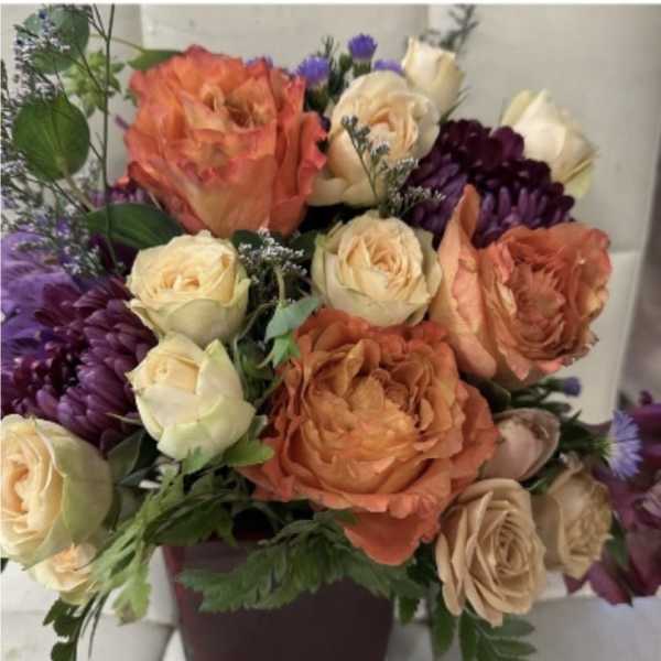 Arrangement of orange and cream roses with purple mums in a dark square vase