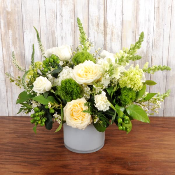Low vase arrangement of cream roses, white flowers, and green accents in a frosted cylinder container.