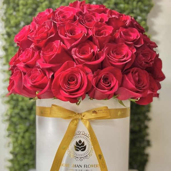 Round box of tightly packed red roses with a gold ribbon on a white hatbox.