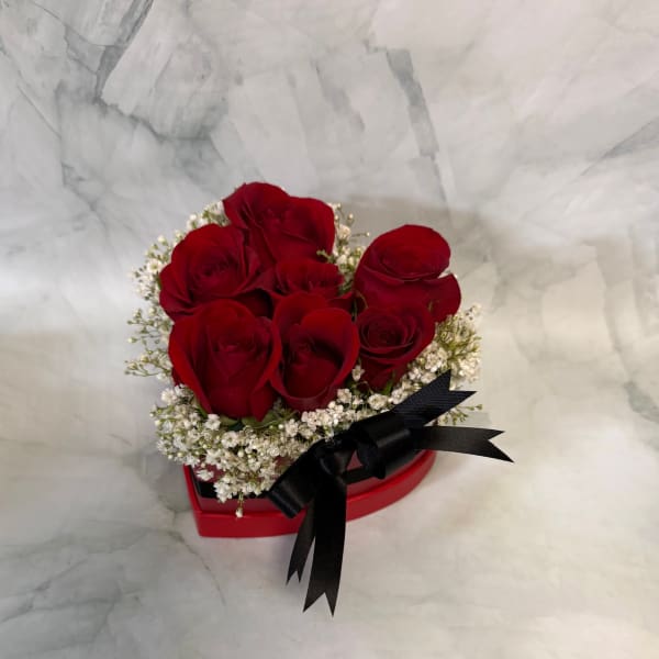 Red roses arranged in a heart-shaped box with white filler flowers and a black ribbon