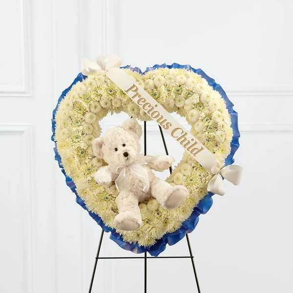 Heart-shaped white flower wreath with a teddy bear and ribbon on a stand
