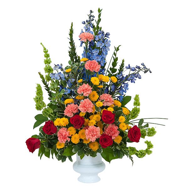Tall mixed arrangement with red roses, pink carnations, yellow mums, and blue delphinium in a white urn.