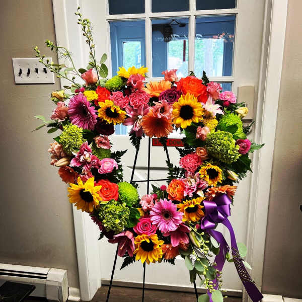 Heart-shaped floral wreath on a stand with bright mixed blooms and a purple ribbon
