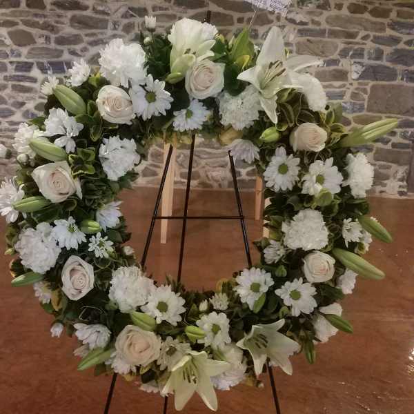 White floral wreath on a stand with lilies, roses, and daisies