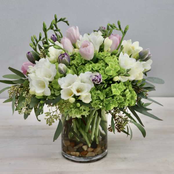 Compact arrangement of pink tulips, white freesias, and green hydrangeas in a clear glass vase with stones.