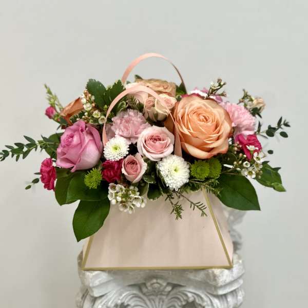 Pastel pink and peach flower arrangement with roses and mums in a blush gift bag-style container.