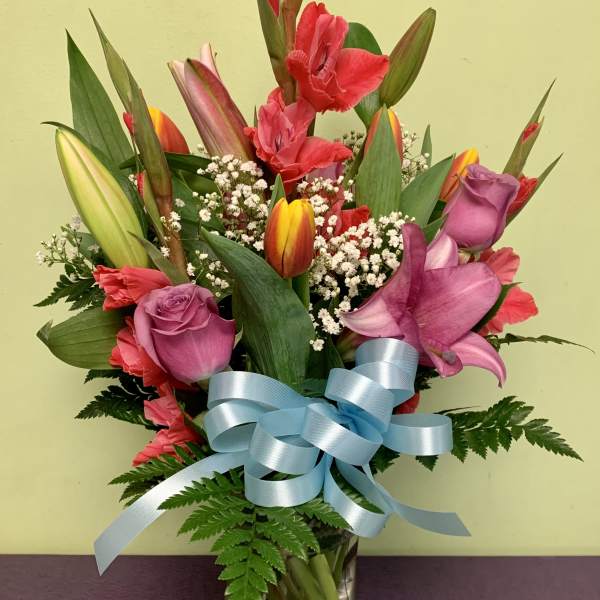 Mixed bouquet of tulips, roses, and lilies in a glass vase with a blue ribbon