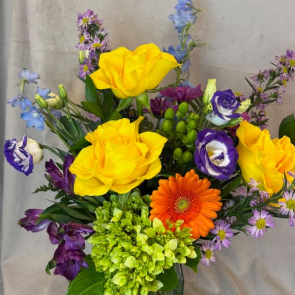 Mixed bouquet with yellow roses, orange gerbera daisy, and purple blooms in a glass vase