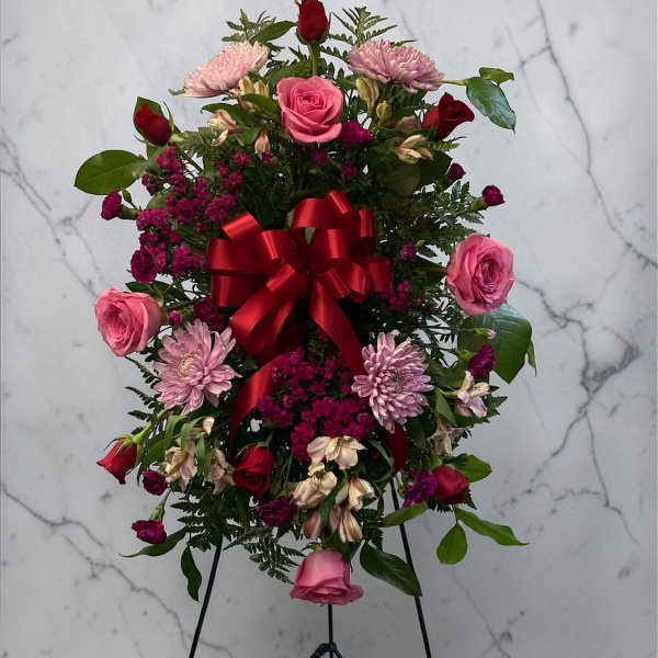 Standing spray of pink and red flowers with a red bow on a black easel.
