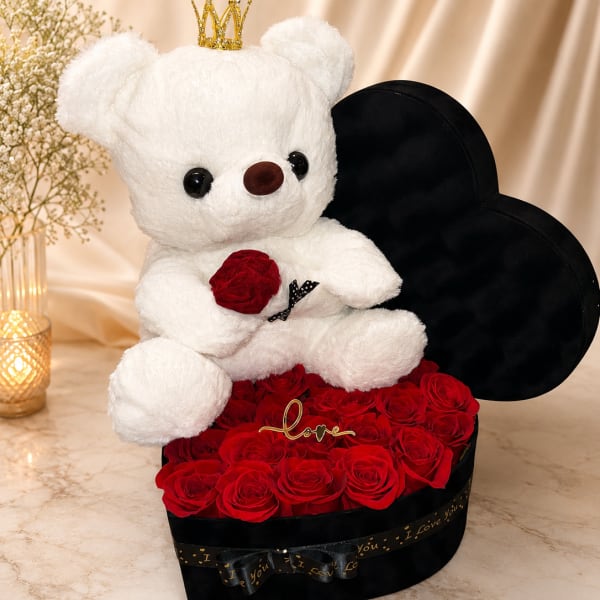 White teddy bear with a crown sitting on red roses in a black heart box