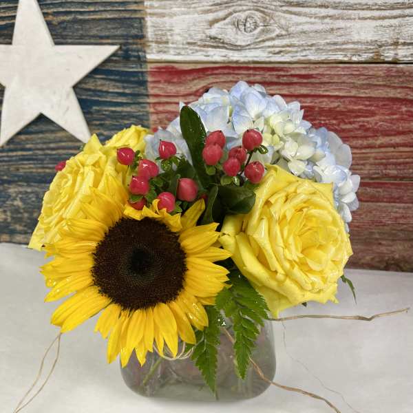 Bouquet of yellow roses, a sunflower, and blue hydrangeas in a glass vase