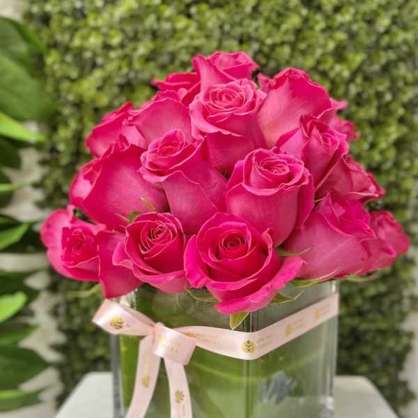 Compact arrangement of bright pink roses in a square glass vase with a pale pink ribbon bow.