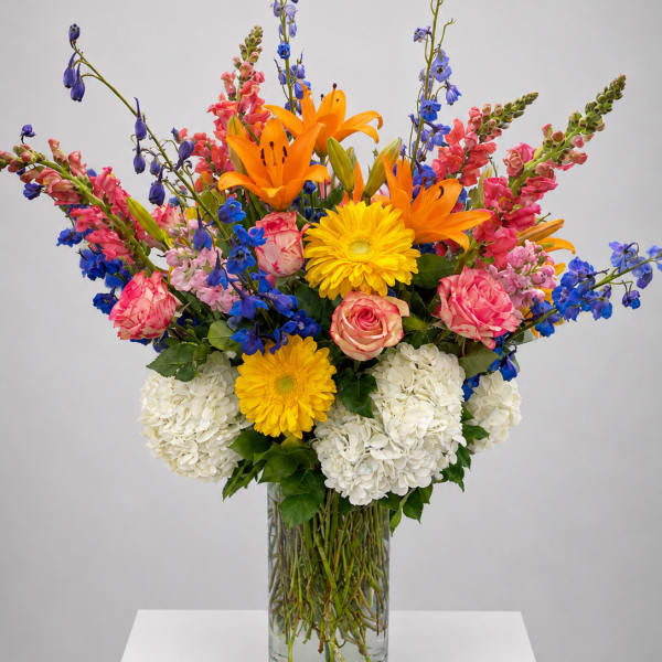 Tall mixed bouquet in a clear glass vase with orange lilies, yellow daisies, pink roses, and blue flowers