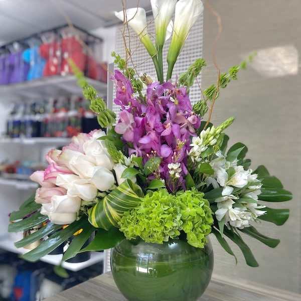 Tall arrangement of white calla lilies, roses, orchids and green hydrangea in a round green glass vase with bold leaves