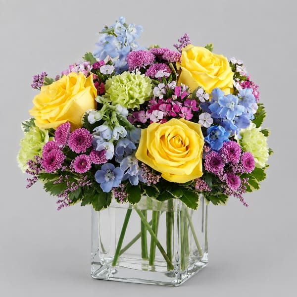 Mixed bouquet of yellow roses, blue flowers, and purple blooms in a clear glass vase