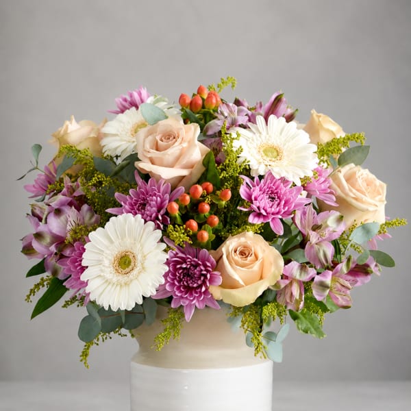 Mixed bouquet of roses, gerbera daisies, and purple blooms in a white vase