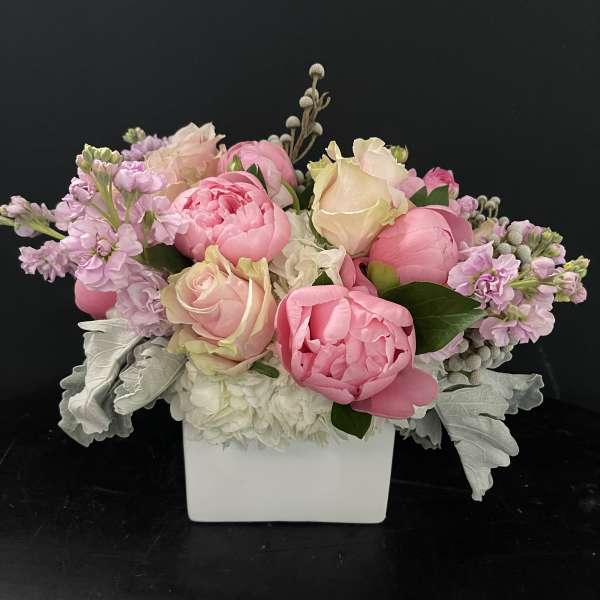 Low arrangement of pink peonies and blush roses in a white square vase with soft white flowers.