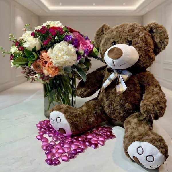 Bouquet of mixed flowers beside a teddy bear with a ribbon and pink heart confetti