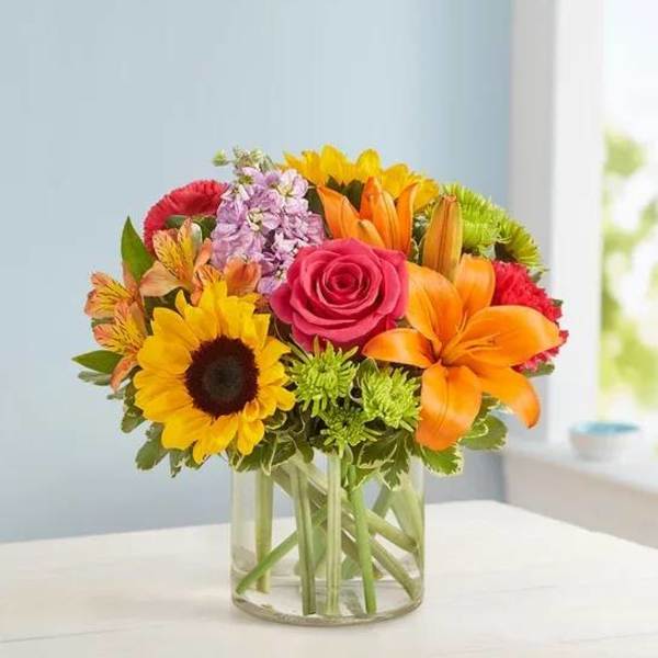 Bright mixed bouquet in a clear glass vase with a sunflower, rose, and lilies.