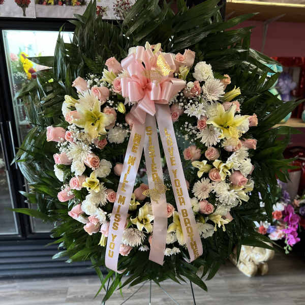 Standing wreath of pink roses, yellow lilies, and white mums with pink bow on an easel