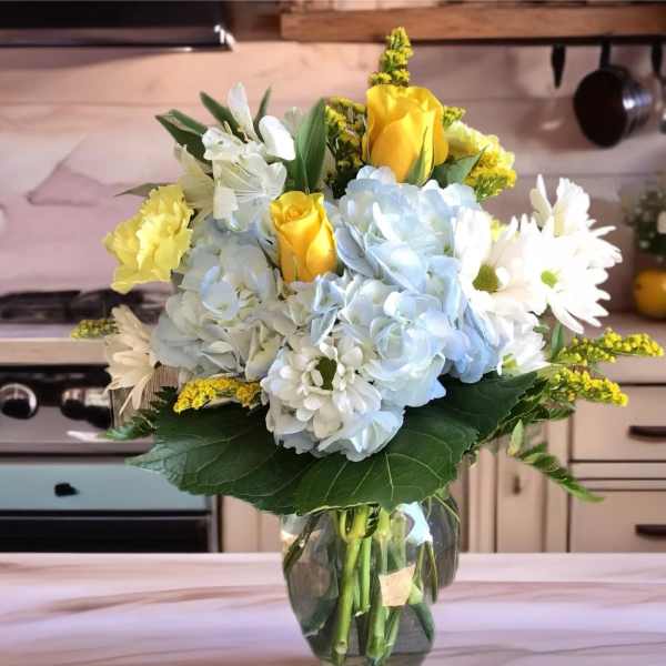 Bouquet of yellow roses, blue hydrangeas, and white daisies in a glass vase