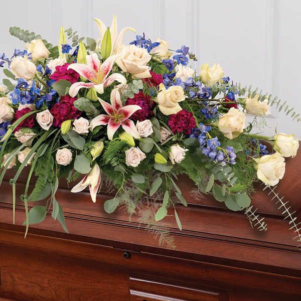 Large mixed flower casket spray with lilies, roses, and blue blooms on a wooden casket