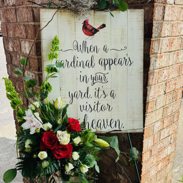Standing floral easel with red, white, and green flowers beside a memorial sign