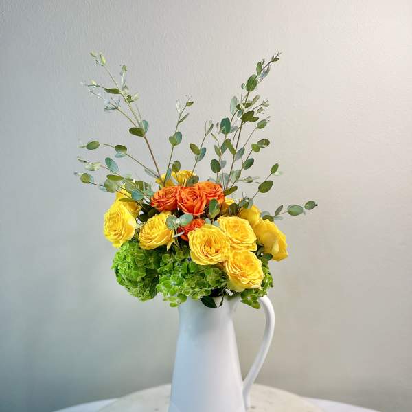 Yellow and orange roses in a white pitcher vase with airy greenery