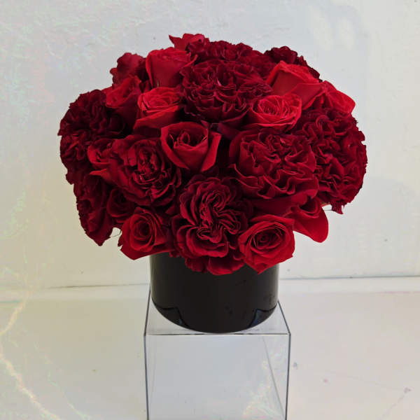 Compact arrangement of red roses and carnations in a black vase on a clear cube stand