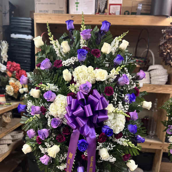 Tall standing sympathy spray of purple, white, and burgundy flowers with a large purple bow on an easel