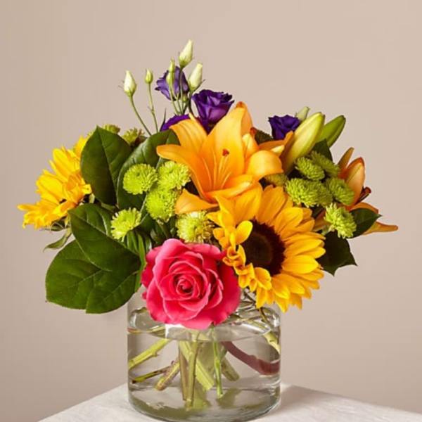 Mixed bouquet in a clear glass vase with pink rose, lilies, and sunflowers