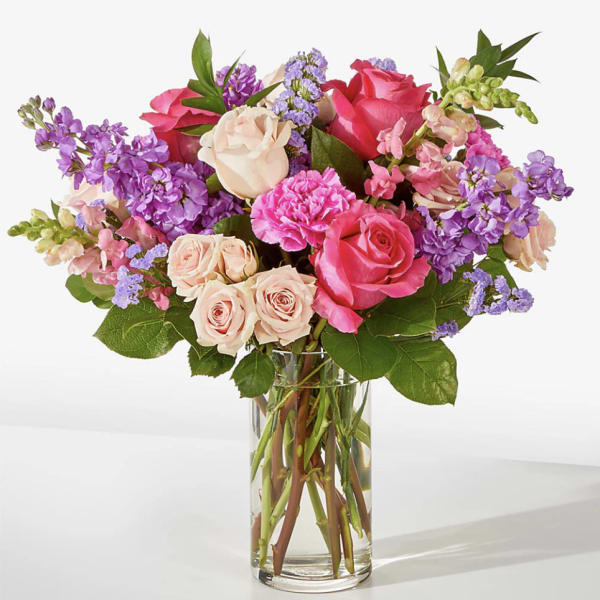 Mixed bouquet of pink roses, carnations, and purple flowers arranged in a clear glass vase